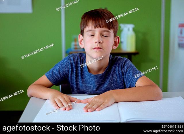 Blind caucasian schoolboy sitting in classroom with eyes closed reading braille book with fingers