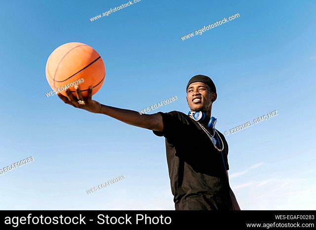 Young man holding basketball while standing against clear blue sky during sunny day