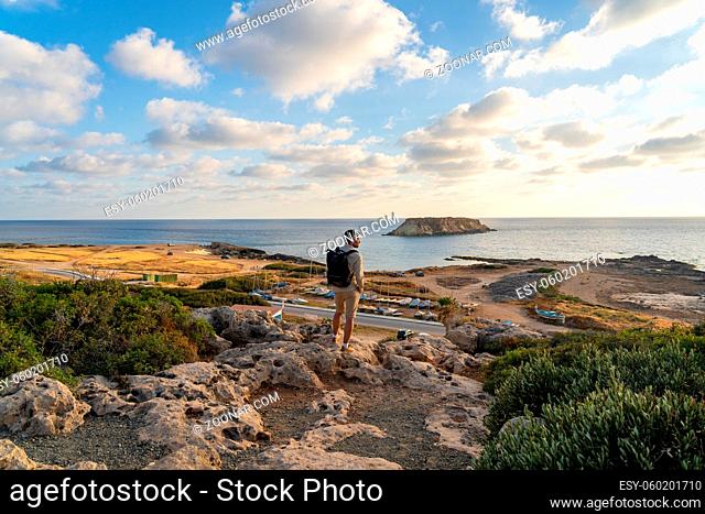 Sea sunset view. Man with backpack on rocks with beautiful view of Yeronisos Island near coast of Agios Georgios Pegeias