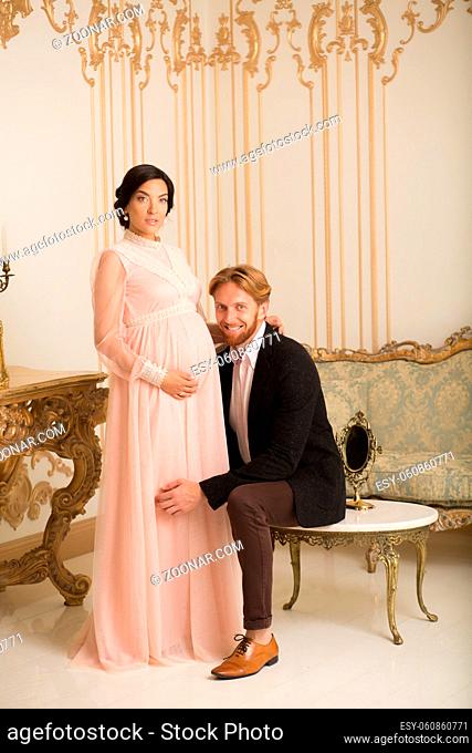Picture of happy rich couple expecting a baby. Brunette pregnant lady in pink dress while husband hugging her. Royalty