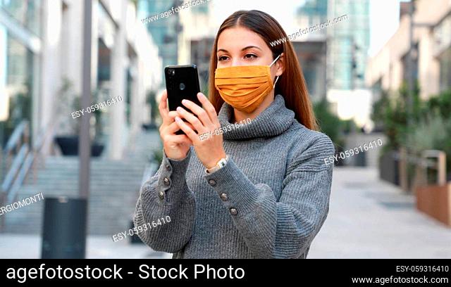 Portrait of young woman standing on street wearing protective mask and messaging with smartphone