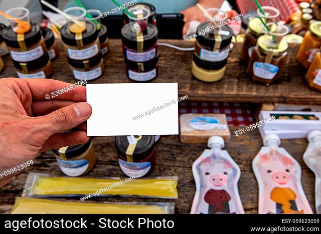 Jams and preserves are seen in the background, displayed on a traditional market stall, with a point of view perspective of a person holding a business card