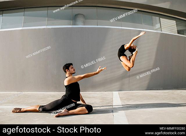 Female dancer doing acrobat while male gymnast practicing stag leap by gray wall