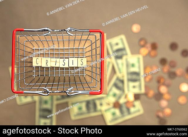 Crisis written with wooden cubes in iron shopping basket with Money coins and bills on the background, Business and Financial concept space for text