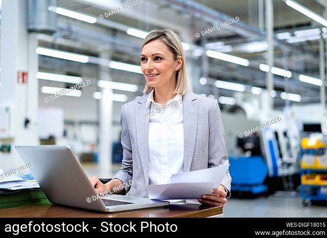 Smiling businesswoman with laptop and documents in factory