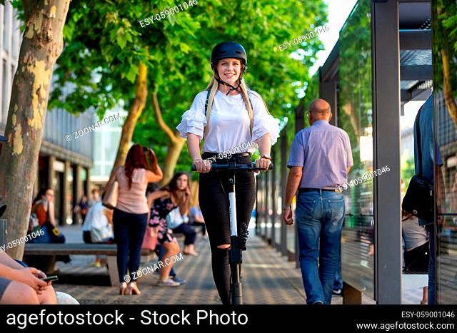 Trendy fashinable teenager, beautiful blonde girl riding public rental electric scooter in urban city environment. Eco-friendly modern public city transport