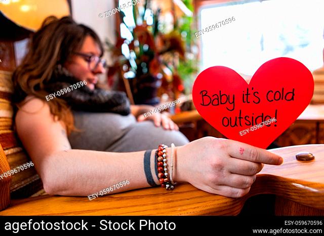 A selective focus view of a heavily pregnant woman holding a red heart message saying baby its cold outside, preparing for childbirth at valentine