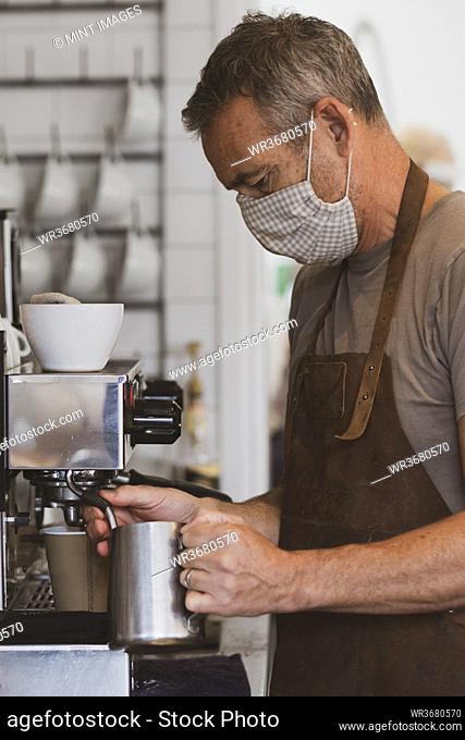 Male barista wearing brown apron and face mask working in a cafe, frothing milk