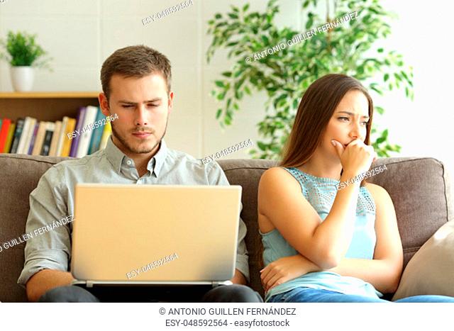 Husband using a laptop on line ignoring to his sad wife sitting on a sofa at home