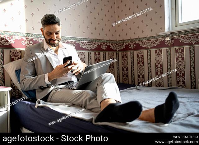 Smiling male professional using smart phone while sitting on bed at home
