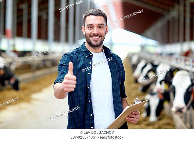 agriculture industry, farming, people and animal husbandry concept - happy smiling young man or farmer with clipboard and cows in cowshed on dairy farm showing...