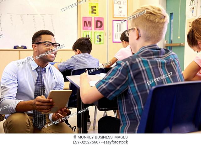 Teacher using tablet computer kneeling beside pupil’s desk