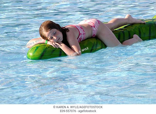 10-year-old girl in pink-and-white two-piece swimsuit lying contentedly on a floating plastic animal in a public swimming pool, in sunshine -- Bloomington