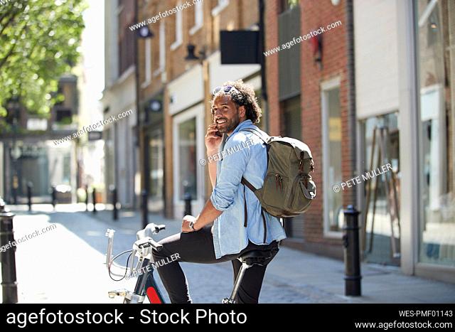 Portrait of smiling young man on the phone with rental bike and backpack in the city, London, UK