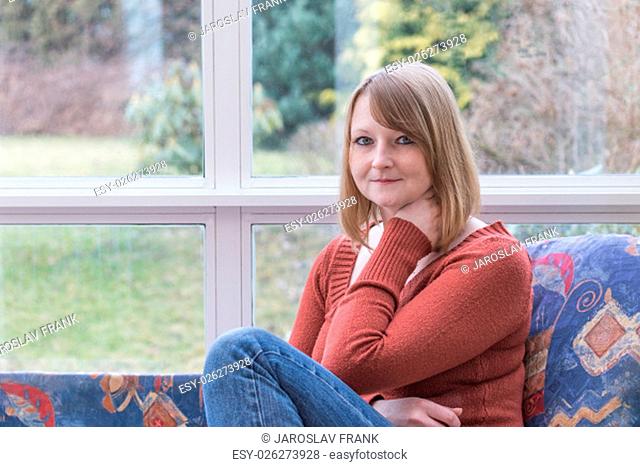 Toothless smiling young woman dressed in a brown sweater is sitting on the sofa in conservatory and she is looking at the camera