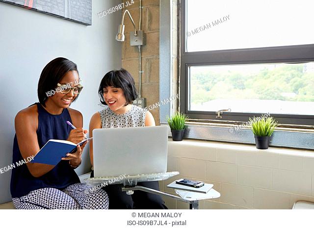 Woman working on laptop, on laptop stand, colleague sitting beside her writing in notebook