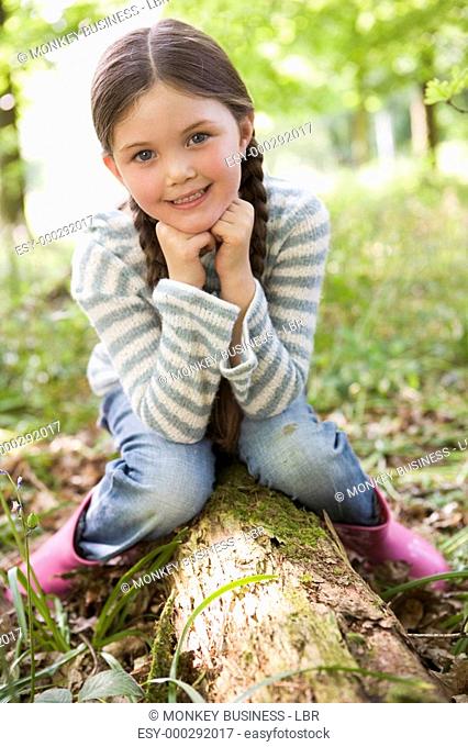 Young girl outdoors in woods sitting on log smiling