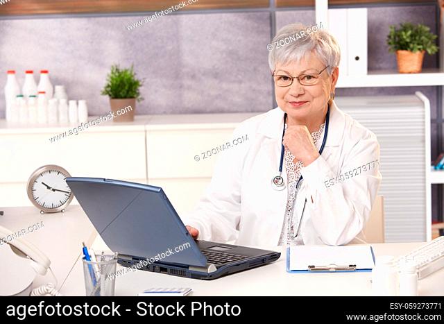 Portrait of senior doctor in office, sitting at desk, looking at camera, smiling