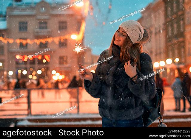 Beautiful girl holding a sparkler enjoys Christmas mood in old European city on outdoor skating rink background with snow and cozy bokeh lights