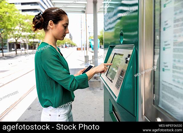 Woman with smart phone using ticket machine at tram stop