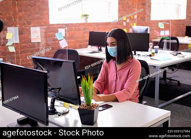 Mixed race businesswoman sitting in office in front of computer wearing face mask