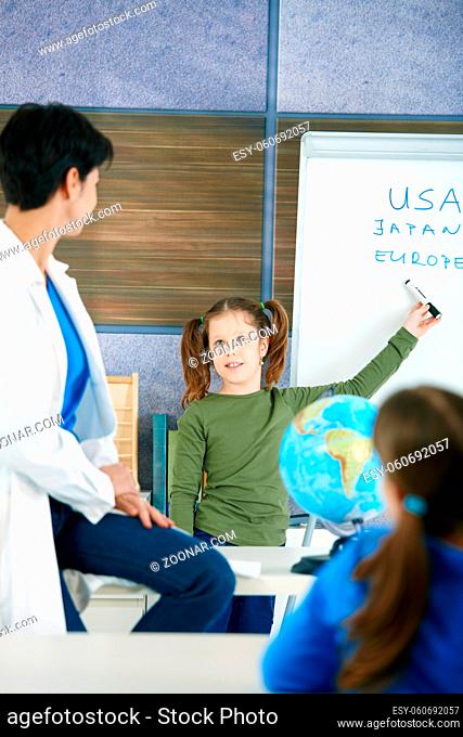 Elementary age schoolgirl at whiteboard answering to teacher in geography lesson