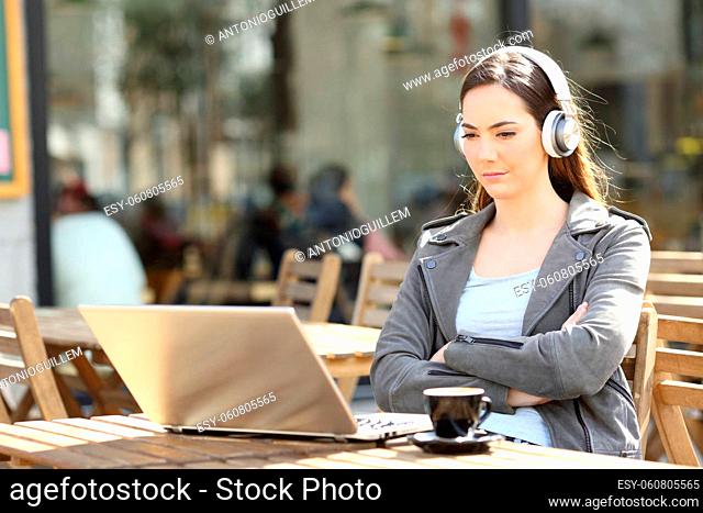 Serious girl crossing arms watching media content on laptop with headphones on a restaurant terrace