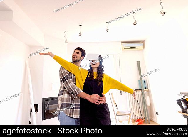 Man embracing happy girlfriend wearing VR glasses in new living room