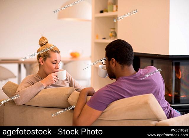 Young romantic multiethnic couple sitting on sofa in front of fireplace at home, looking at each other, talking and drinking coffee