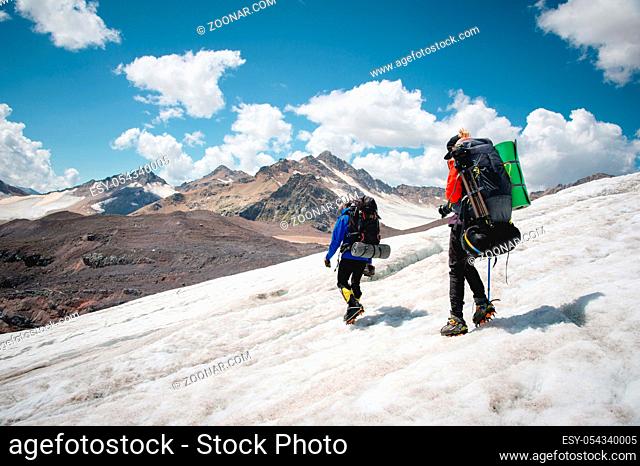 Two tourists, a man and a woman with backpacks and crampons on their feet walk along the glacier against the background of the mountains of the sky and clouds
