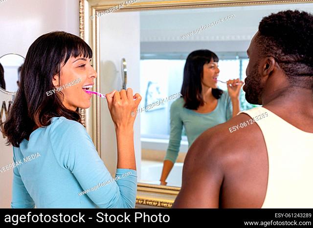 Happy multiracial young couple brushing teeth together in front of mirror at home