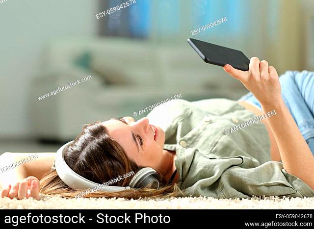 Happy woman listening to music with smart phone and headphones lying on a carpet at home