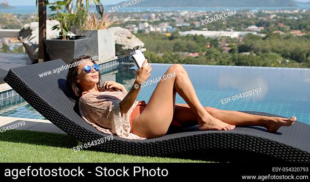 Side view of brunette in beachwear spending time on terrace of luxury hotel resort chilling on deckchair near pool and taking selfie with smartphone