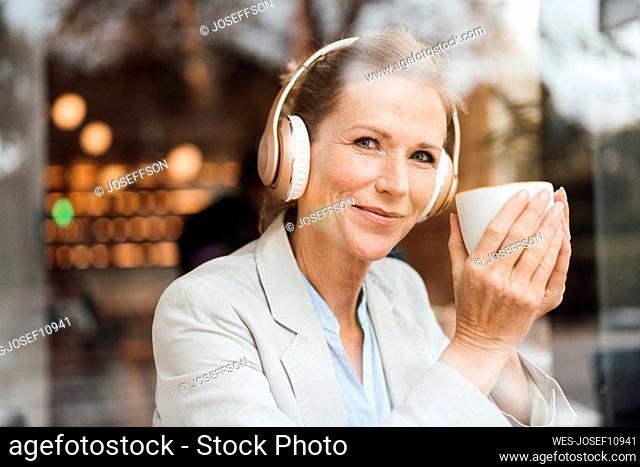 Smiling businesswoman holding coffee cup enjoying music listening through wireless headphones in cafe