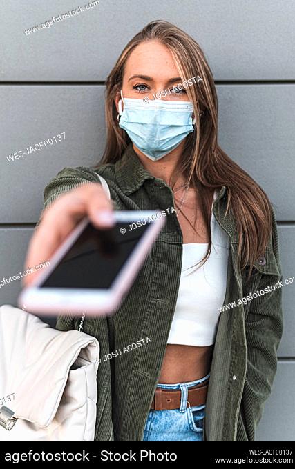Young woman wearing face mask giving smart phone while standing against wall