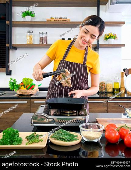 Young asian woman dressed in an apron use tongs to tong the steak from the hot pan. Morning atmosphere in a modern kitchen