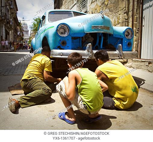 Man man repairs a broken down car in the street while another man and a boy look on in Havana, Cuba