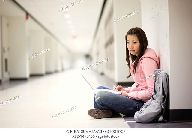 Portrait of a young Asian woman using her laptop computer while sitting on floor in school hallway