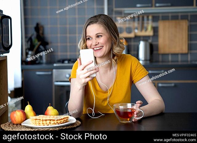 Happy woman with smartphone and earbuds sitting at kitchen table at home