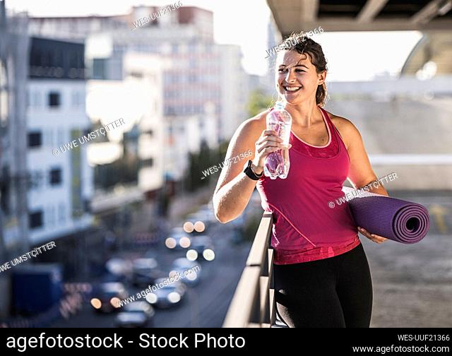 Smiling young woman holding mat looking away while drinking water by railing