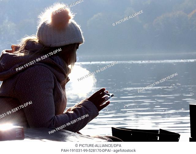 31 October 2019, North Rhine-Westphalia, Essen: Despite the morning cold at Lake Baldeney, a young woman sits on the terrace of an excursion restaurant in the...
