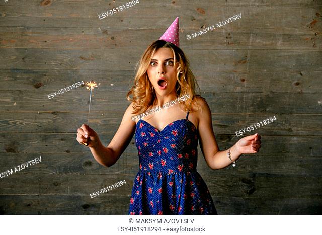 Attractive cheerful young woman looks pleasantly wondered, widely open mouth , celebrating party, holds the sparkler fire, dressed up in blue dress, festive cap
