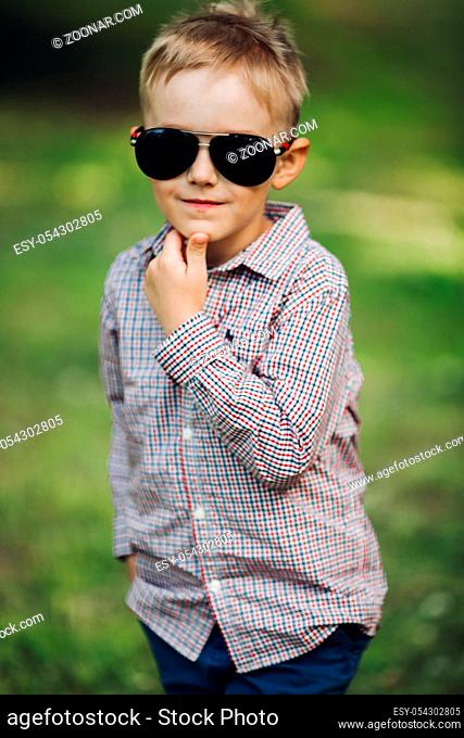 Portrait of stylish little boy, wearing in checkered shirt and cool sunglasses posing in park in summertime. Handsome boy holding one hand in pocket and other...