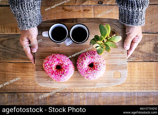 above view of pair of caucasian female hands taking a wood plate with two black italian coffee and a couple of pink sugar donuts, wood table in background