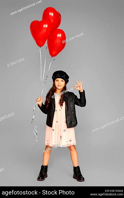 fashionable child girl holding red heart shaped balloons and showing Ok gesture isolated on gray background. valentine's day