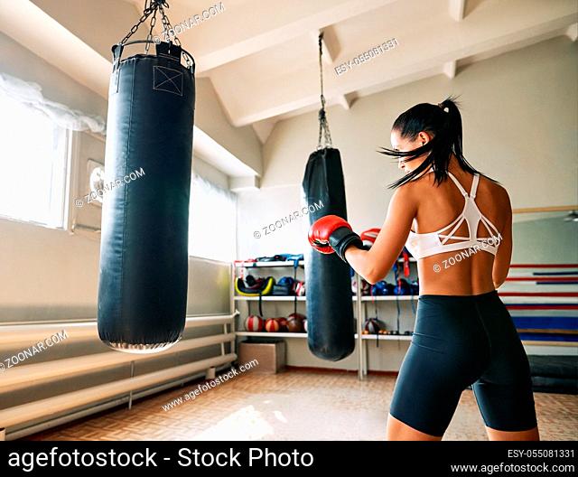 Back view of female boxer hitting a huge punching bag at fitness gym. Woman practicing her punches at a boxing studio