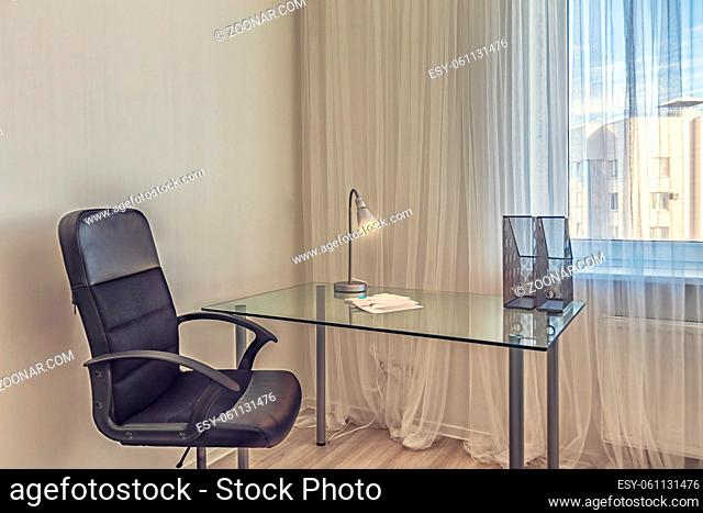 Clean modern home office with transparent glass desk and black armchair near window