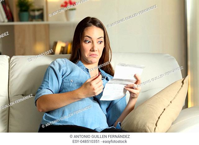 Distrustful pregnant lady reading a leaflet before take a pill sitting on a couch in the living room at home
