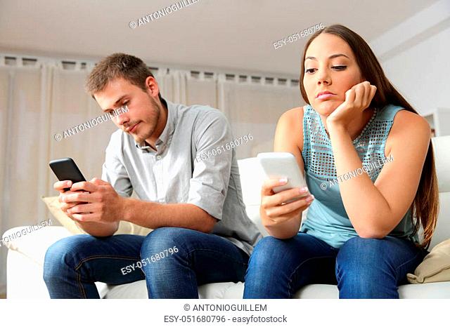Bored couple online with their smart phones sitting on a couch in the living room at home