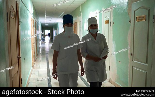 Two masked female nurses in white coats walking through hospital hall looking at camera. Hospital interior in process of repair